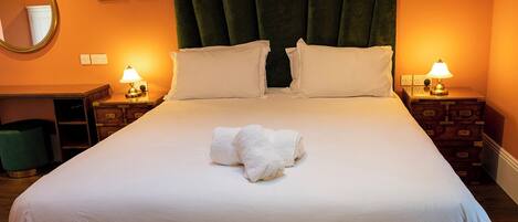Una cama impecablemente tendida con sábanas blancas y dos almohadas, un cabecero tapizado en verde, mesitas de noche con lámparas y un espejo en la pared.