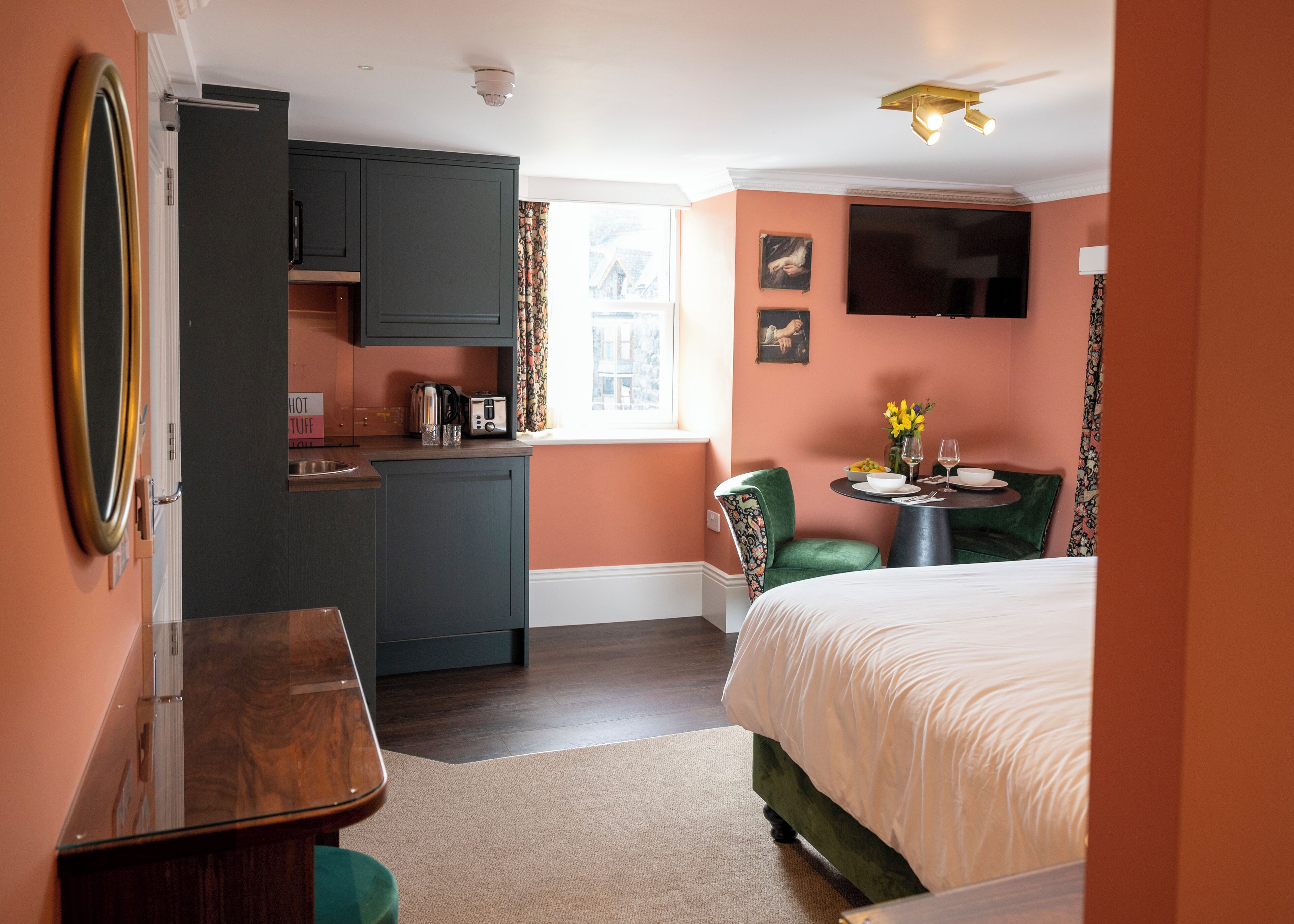 A compact hotel room with a kitchenette, a bed, a small table with a chair, and a TV mounted on the wall.