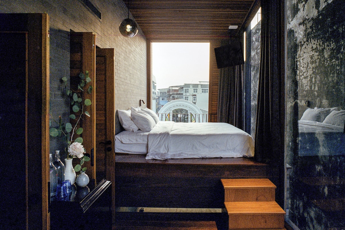 A bedroom with a bed, a nightstand, and a window with a view of buildings.