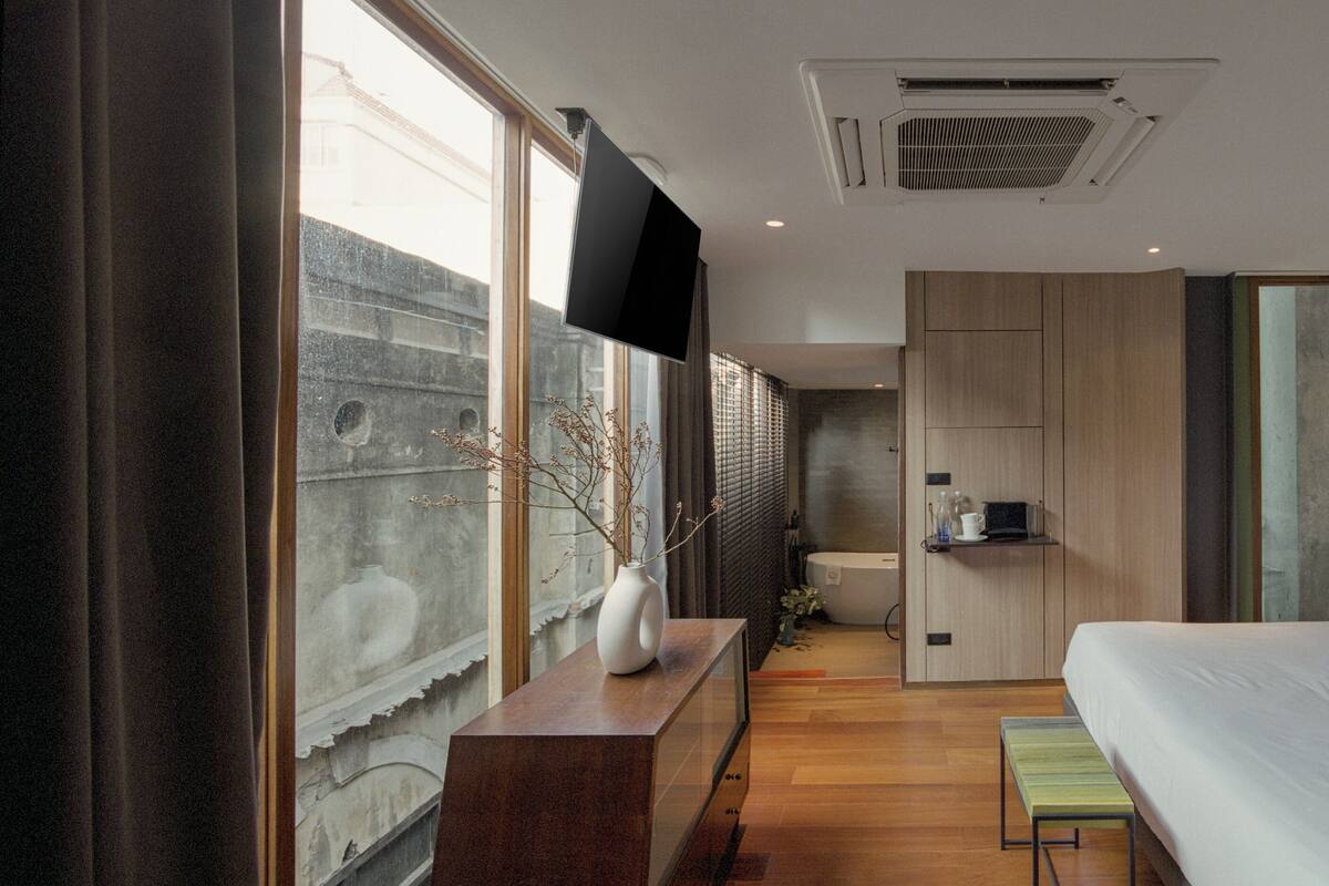 A modern hotel room with a large bed, a TV mounted on the wall, a small bedside table, and a window with a view of a building outside.