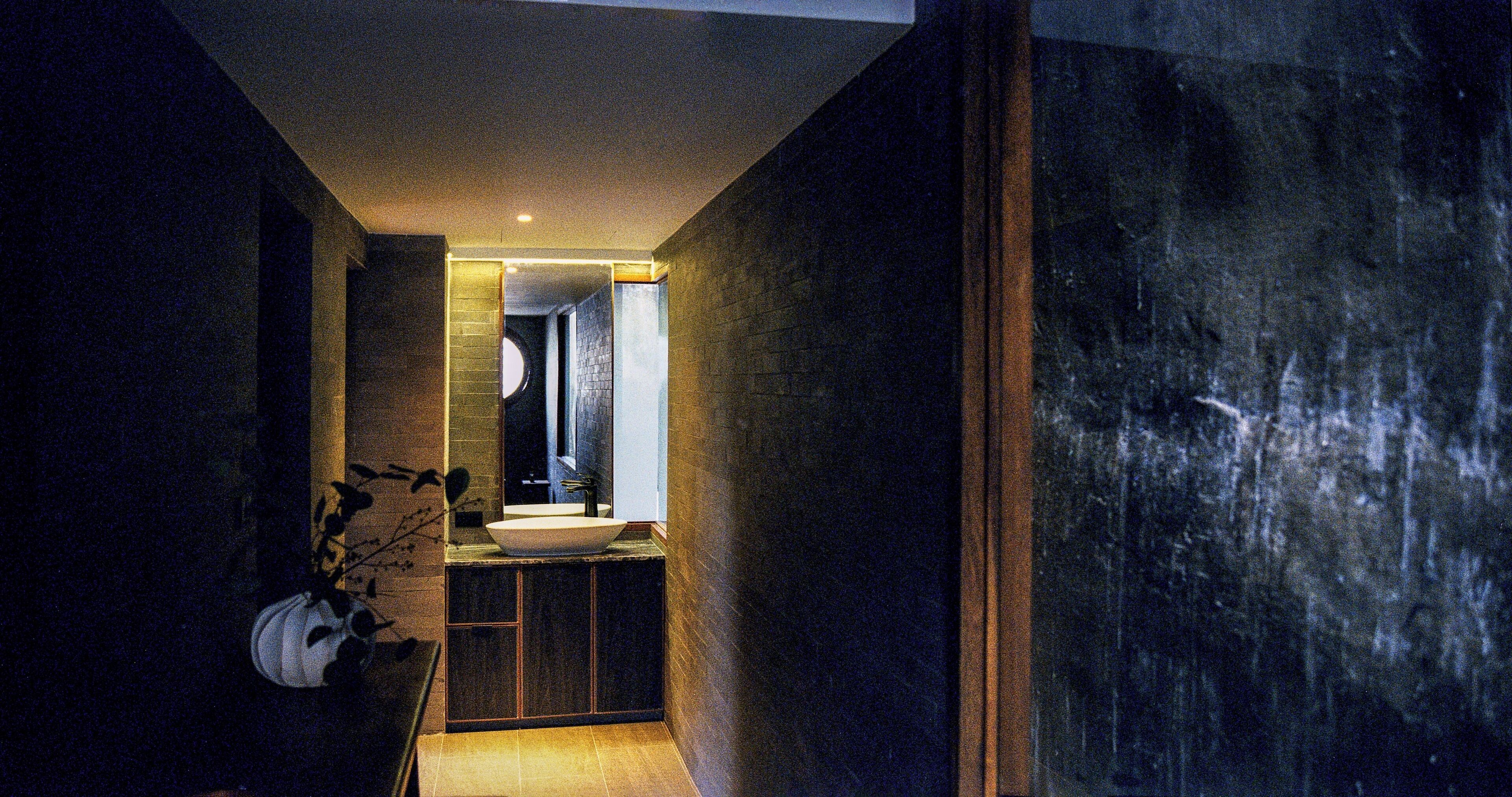 Un pasillo estrecho, tenuemente iluminado, con un baño moderno que incluye un espejo grande, un lavamanos y una pared oscura con textura.