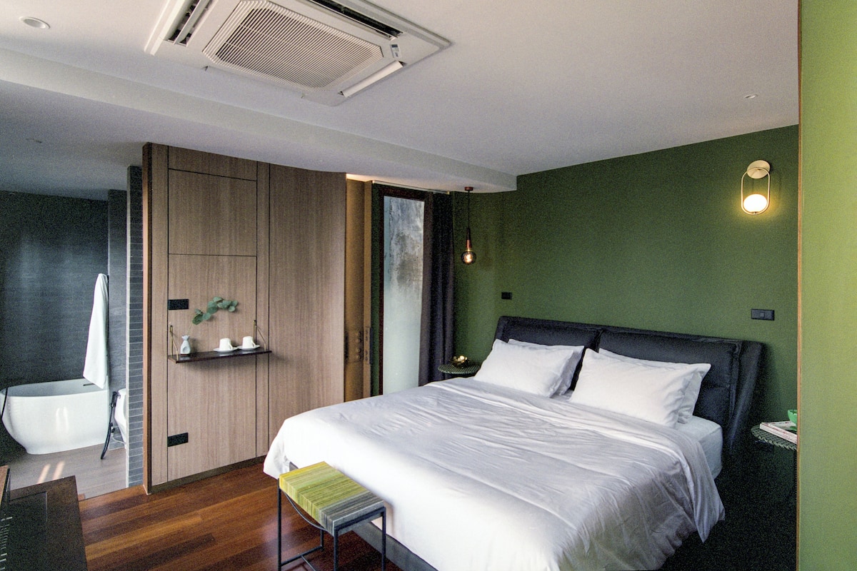 A modern bedroom with a large bed, a bedside table, a small stool, a nightstand, a wall-mounted light, and a ceiling vent.