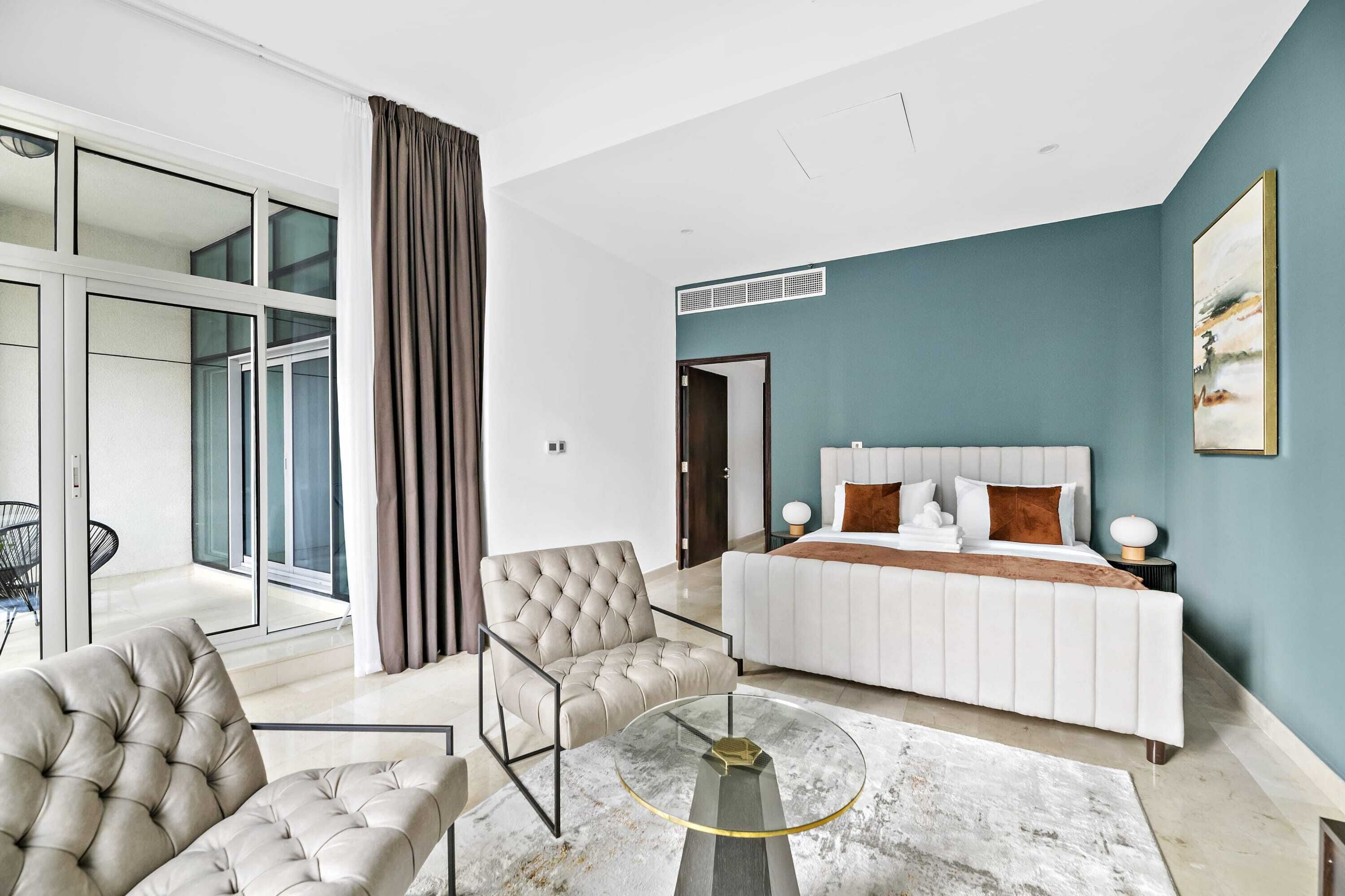 Une chambre moderne avec un grand lit, un fauteuil à capitons, une table basse en verre et une vue sur un balcon.