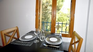 Une table à manger est dressée avec des assiettes, des verres et des couverts, et est placée près d’une fenêtre donnant sur un paysage verdoyant et une rambarde bleue.