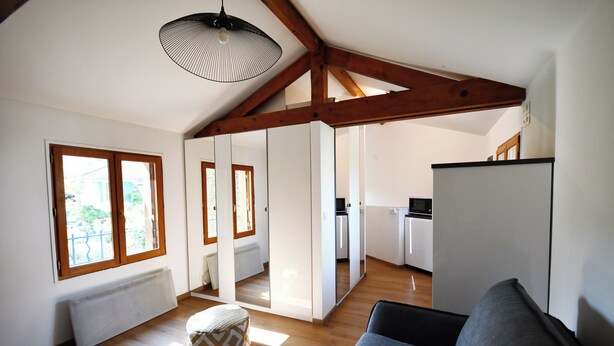 Un vaste espace de vie doté de planchers en bois, d’un canapé gris, d’une petite table et d’un radiateur. De grandes fenêtres laissent entrer la lumière naturelle, et on aperçoit un coin cuisine au fond de la pièce.