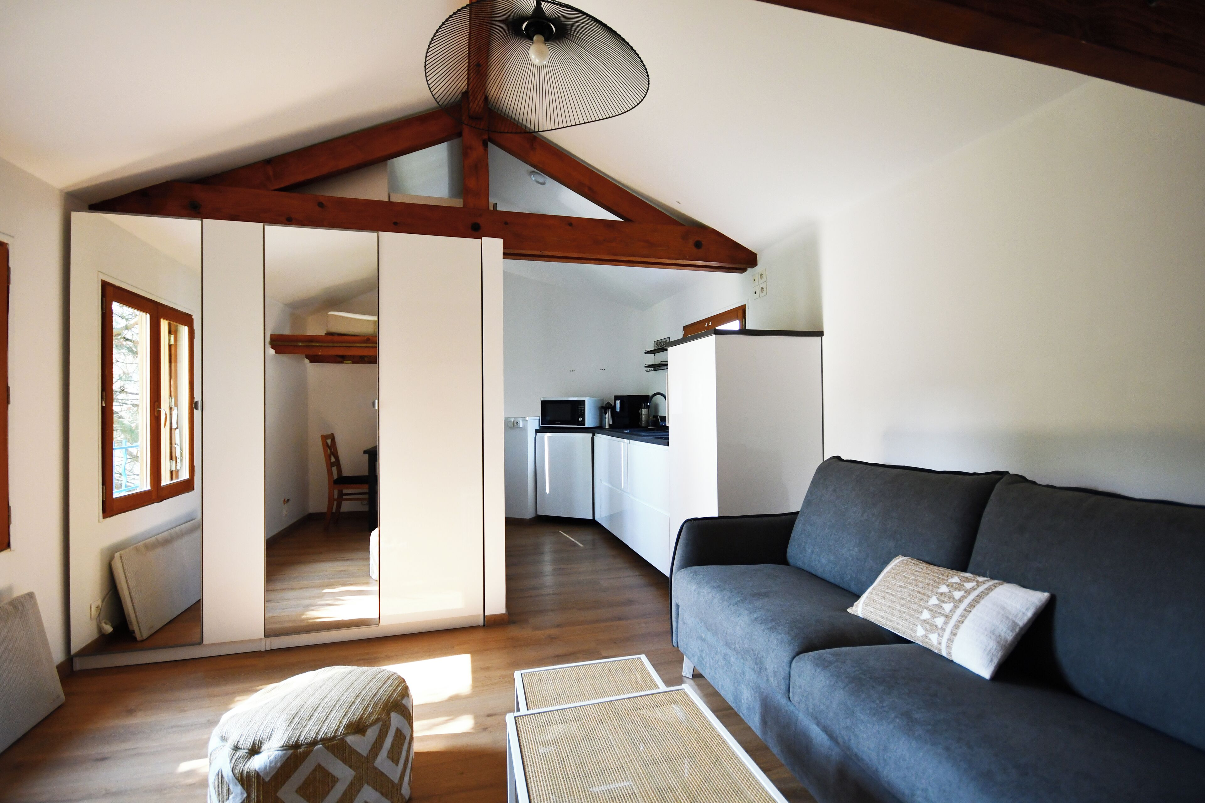 Una sala moderna con un sofá gris, un reposapiés tejido y un ventilador de techo. Se ve una cocina al fondo con armarios blancos y un microondas.