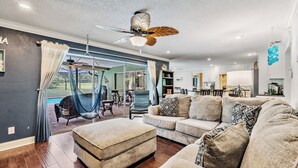 A spacious living room with a sofa, ottoman, and a hammock. There is a ceiling fan, a dining area, and a kitchen in the background.