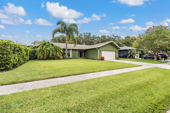 A single-story house with a green exterior, a front lawn, and a driveway leading to a garage. - Clearwater Pool Home - Family Friendly! (Clearwater)
