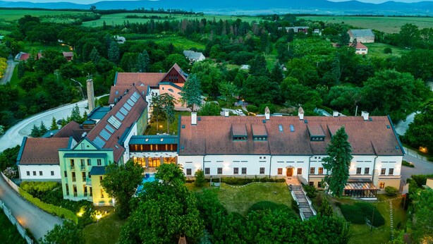 Eine Luftaufnahme eines weitläufigen Anwesens mit mehreren Gebäuden, darunter ein Hauptgebäude mit rotem Ziegeldach und ein kleinerer Anbau mit grüner Dachbegrünung.