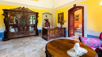 Una habitación con un mueble de madera grande y ornamentado, una mesa redonda de madera con una figurita de un gato blanco, y un sofá colorido con estampado.