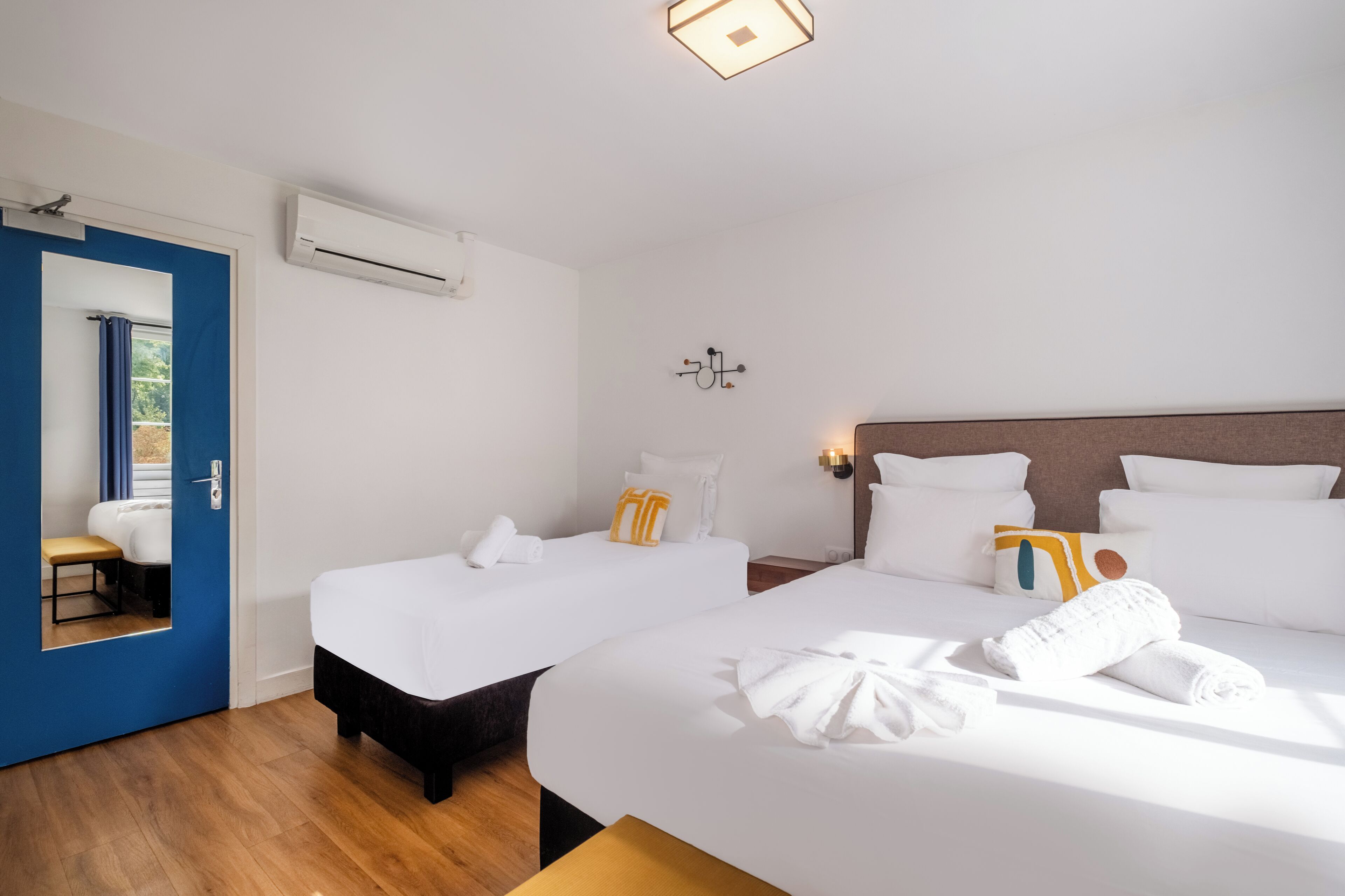A hotel room with two beds, a blue door, an air conditioning unit, and a mirror reflecting a bathroom.