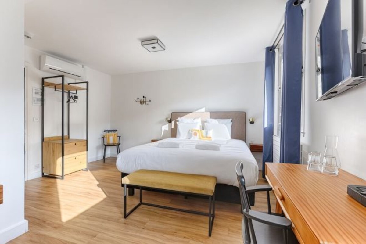 Habitación de hotel con cama, un banco de madera, escritorio, silla, televisor y ventana con cortinas.