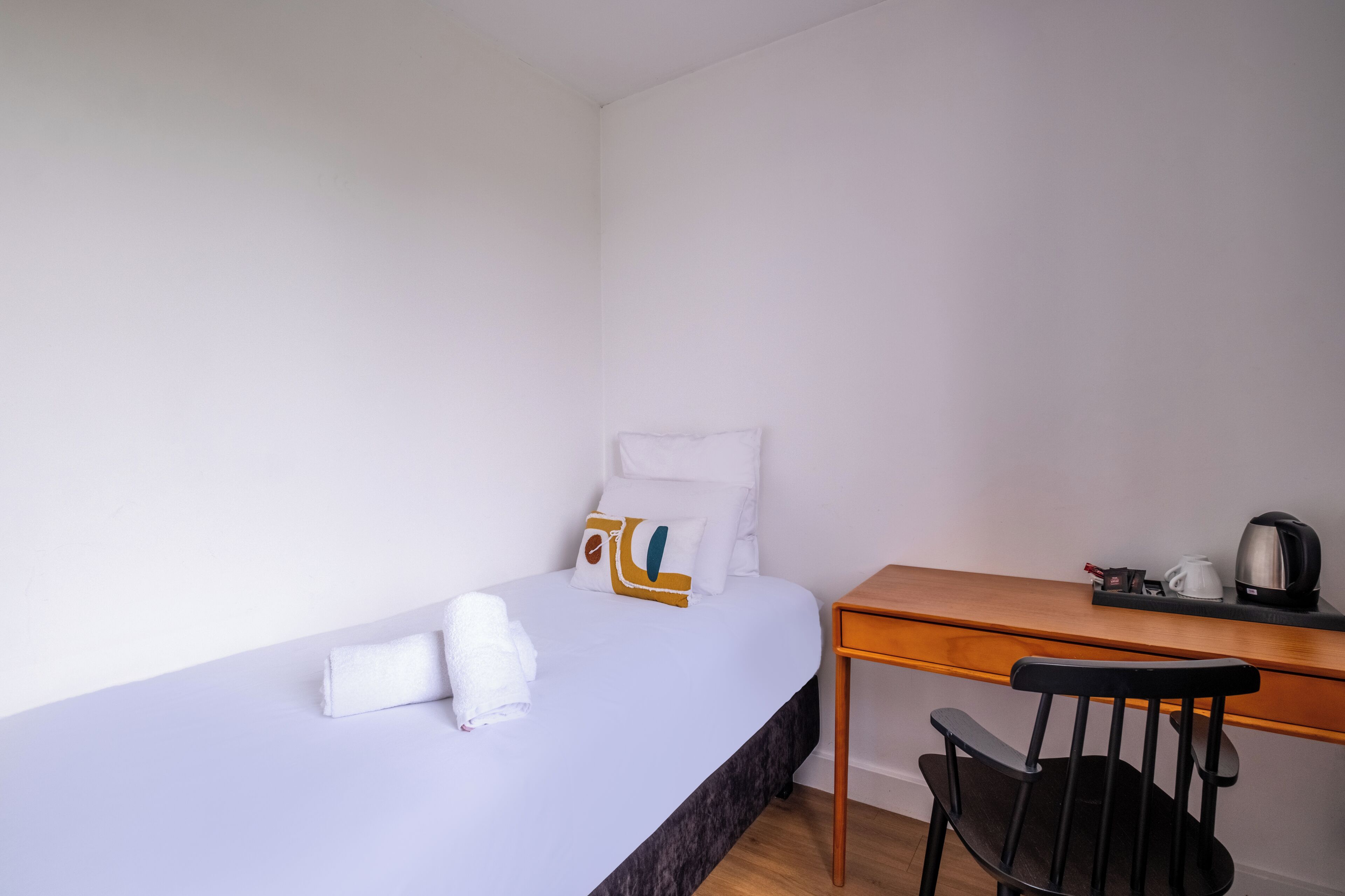 A single bed with white linens and a pillow, a wooden desk with a kettle and a cup, and a black chair.
