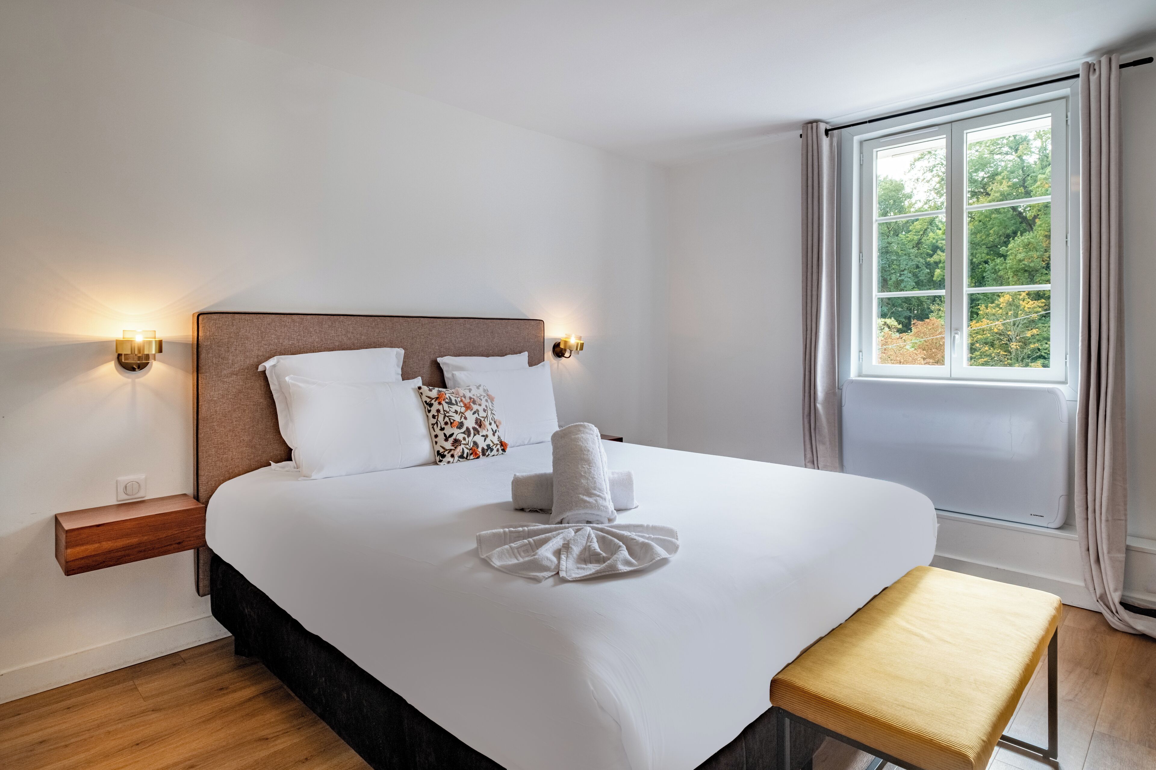 A neatly made bed with a white comforter, pillows, and a folded towel. There is a wooden nightstand with a lamp, a bench, and a window with curtains.