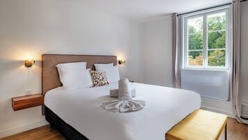 Un lit bien fait, avec une couette blanche, des oreillers et une serviette pliée. On y trouve une table de chevet en bois avec une lampe, un banc et une fenêtre avec des rideaux.