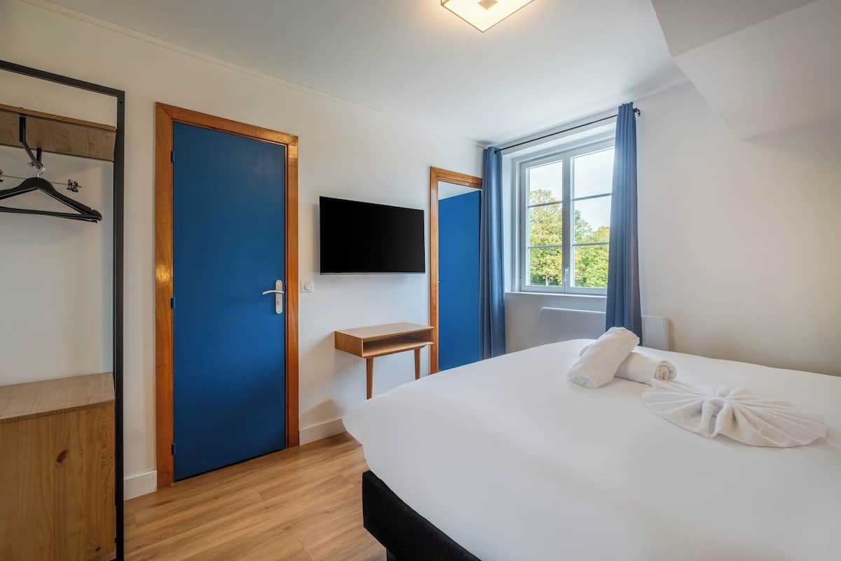 Ein Hotelzimmer mit Bett, einem an der Wand montierten Fernseher, einem Holztisch und einem Fenster mit Vorhängen.