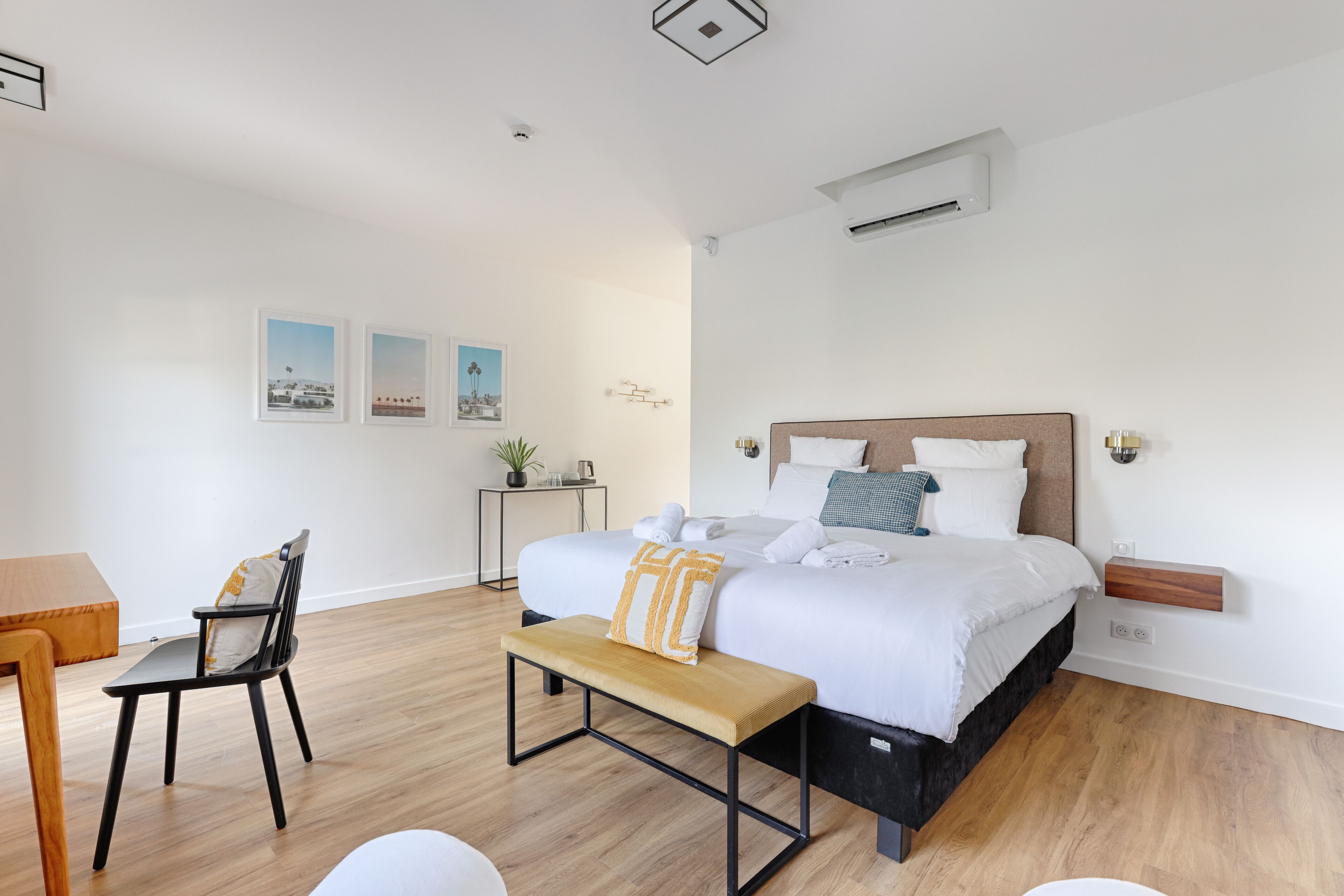 Une chambre moderne avec un plancher en bois, un lit avec une literie blanche, un banc, un bureau et une petite table sur laquelle est posée une plante.