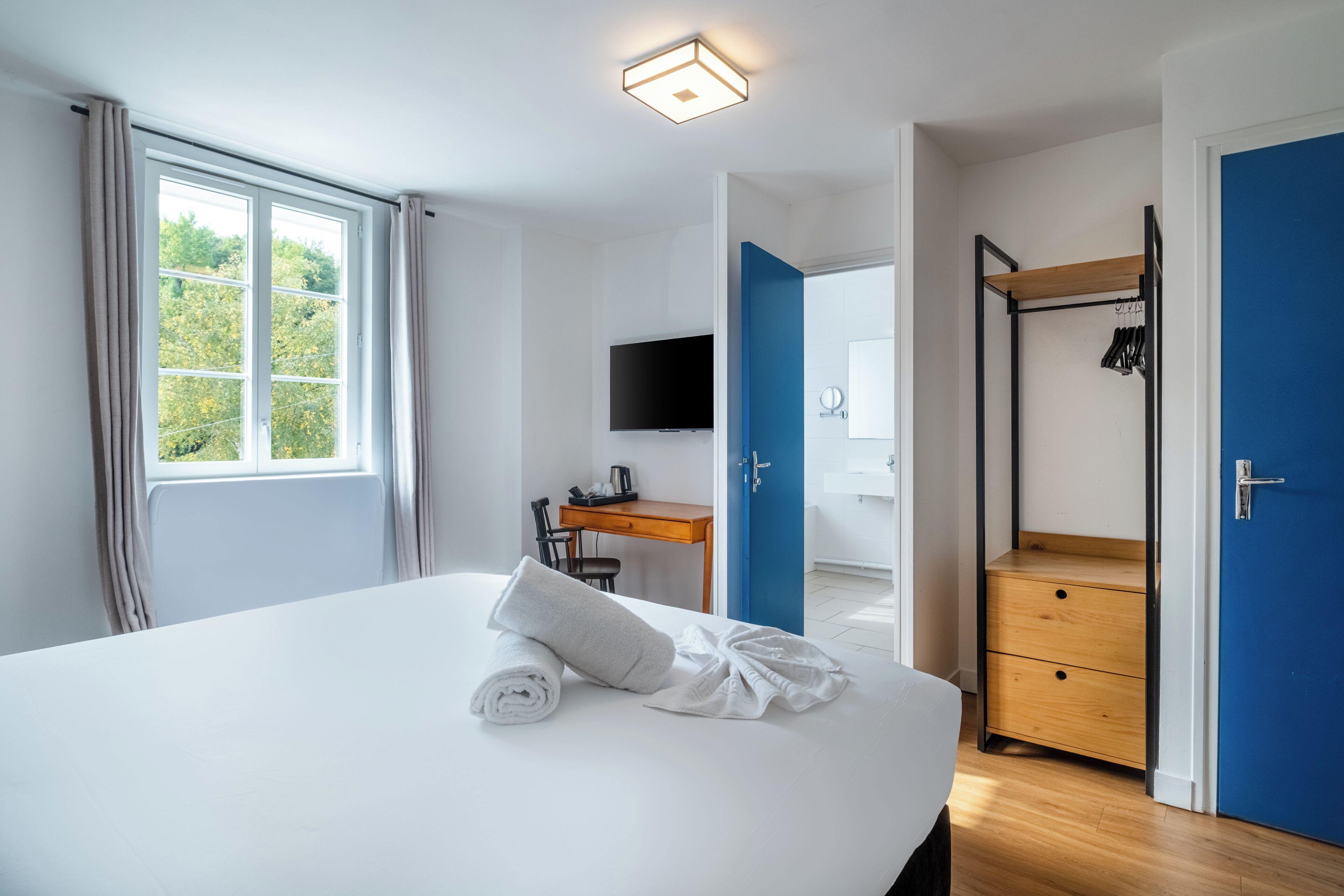 Habitación de hotel con una cama grande, un escritorio con silla, televisión, armario y baño visible a través de una puerta abierta.