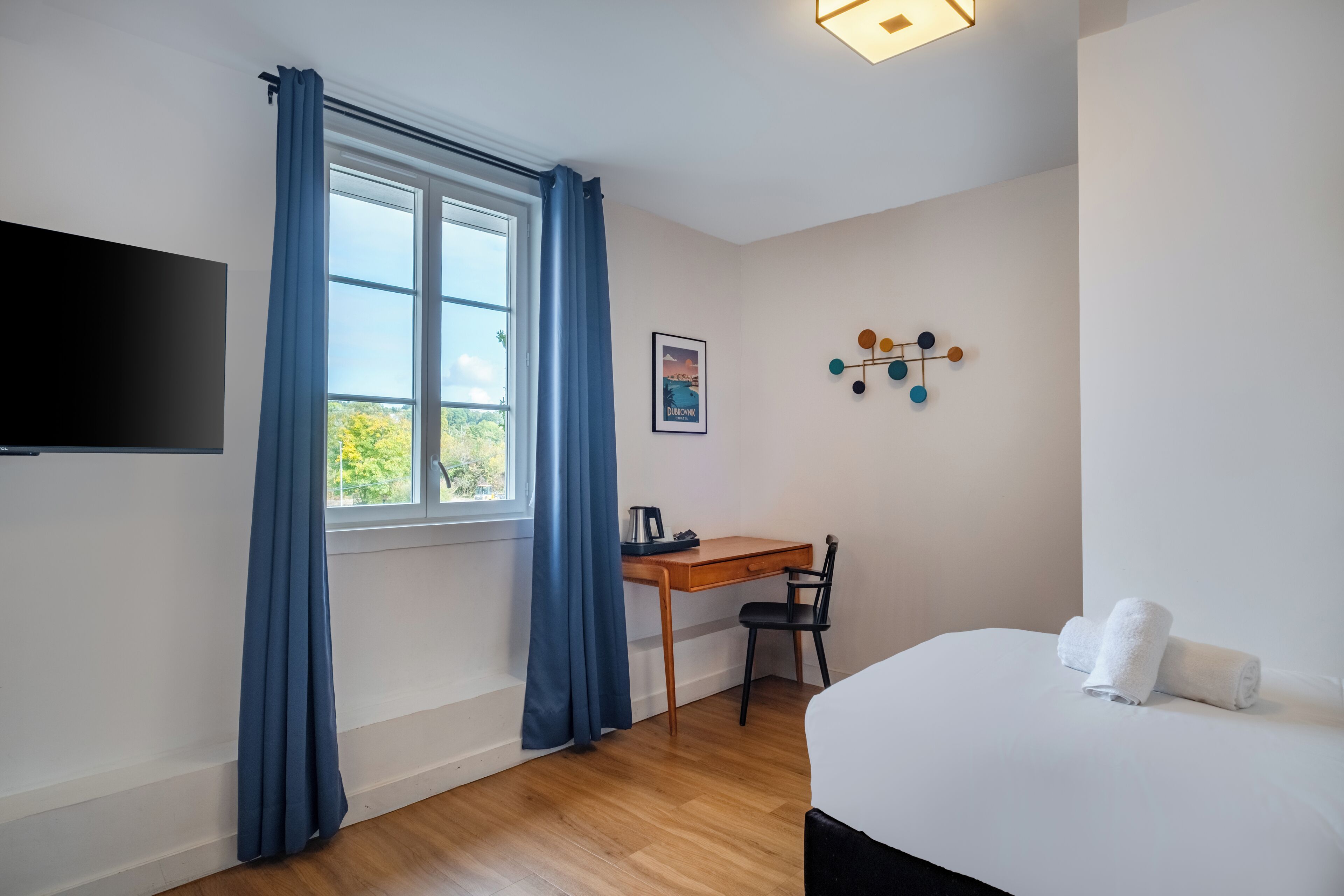 Una habitación de hotel con escritorio de madera, una silla, cama con ropa blanca, televisión de pantalla plana en la pared y ventana con cortinas azules.