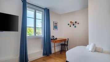 Una habitación de hotel con escritorio de madera, una silla, cama con ropa blanca, televisión de pantalla plana en la pared y ventana con cortinas azules.