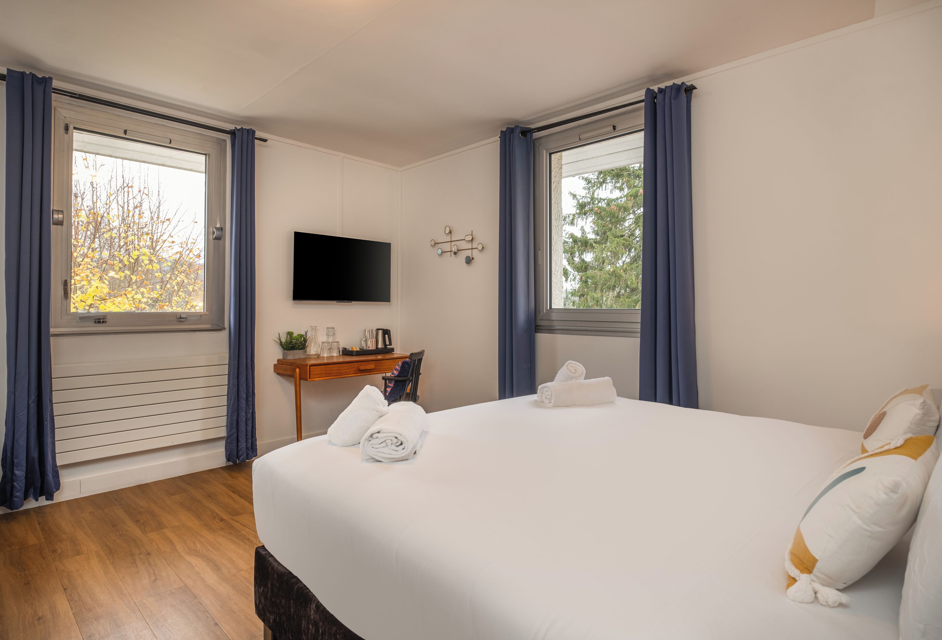 Habitación de hotel con una cama grande, un televisor de pantalla plana montado en la pared, un escritorio pequeño con una planta y dos ventanas con cortinas.