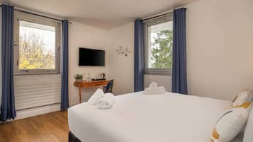 Habitación de hotel con una cama grande, un televisor de pantalla plana montado en la pared, un escritorio pequeño con una planta y dos ventanas con cortinas.
