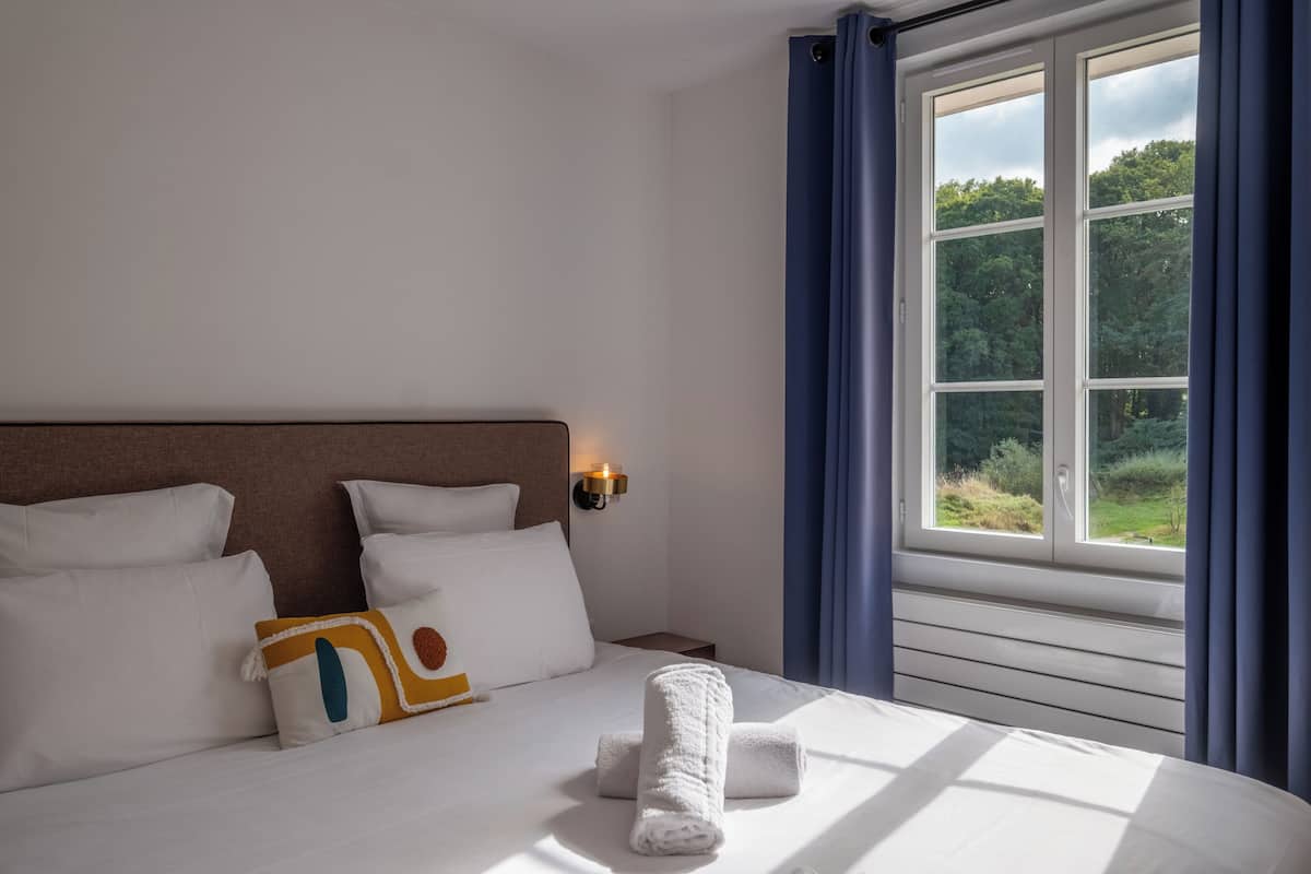 Ein Hotelzimmer mit einem großen Bett, weißer Bettwäsche, einem Kopfteil, einem Nachttisch, einer Wandlampe und einem Fenster mit Vorhängen.