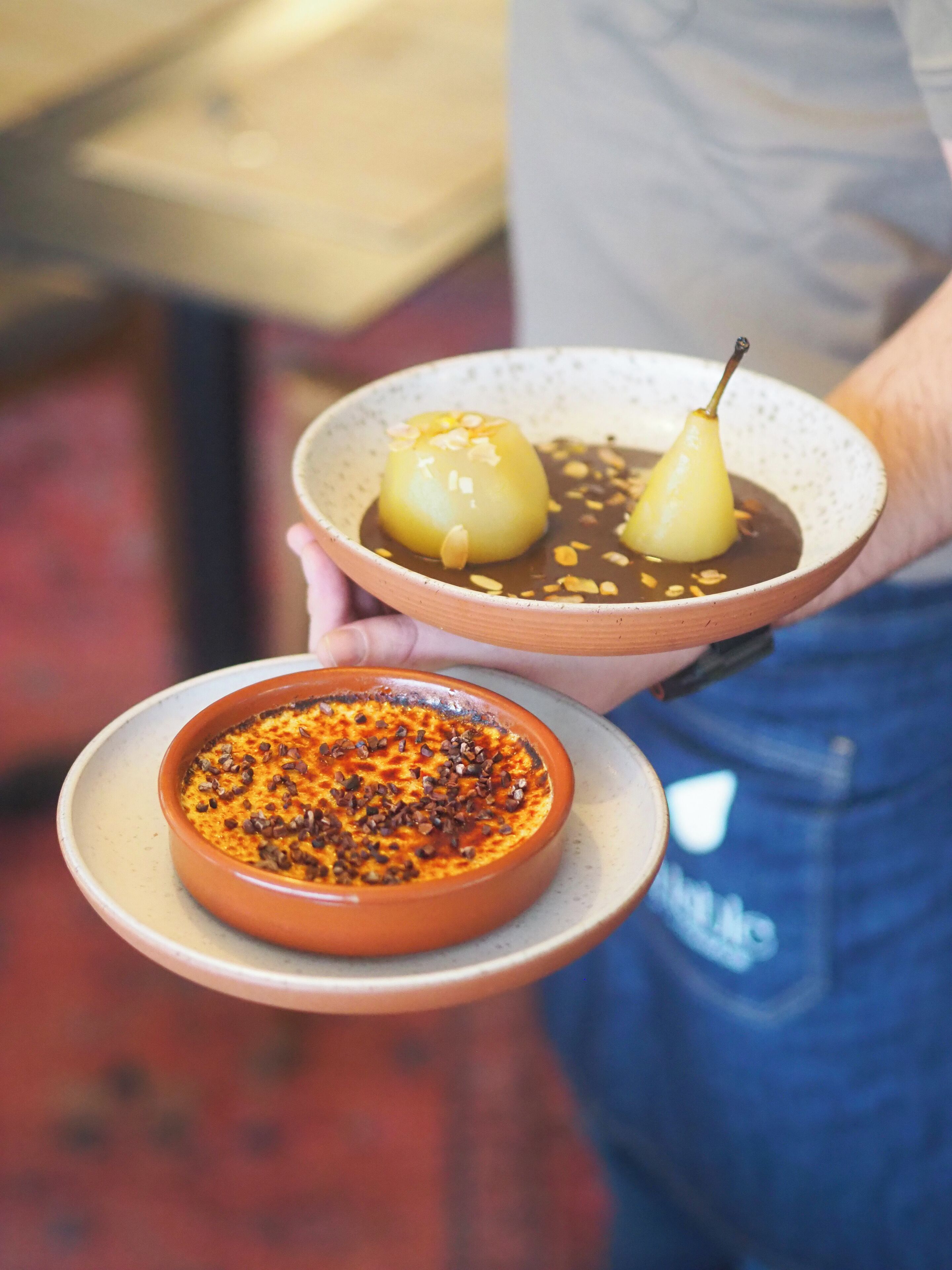 A person holding two dishes, one with a pear and another with a chocolate dessert.