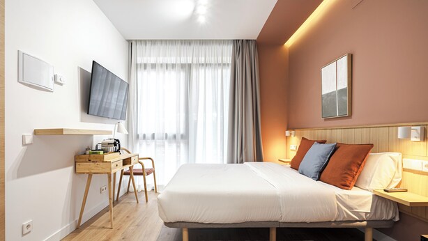 Une chambre d’hôtel moderne équipée d’un lit, d’une table de chevet, d’une télévision, d’un bureau, d’une chaise et d’une grande fenêtre avec des rideaux.