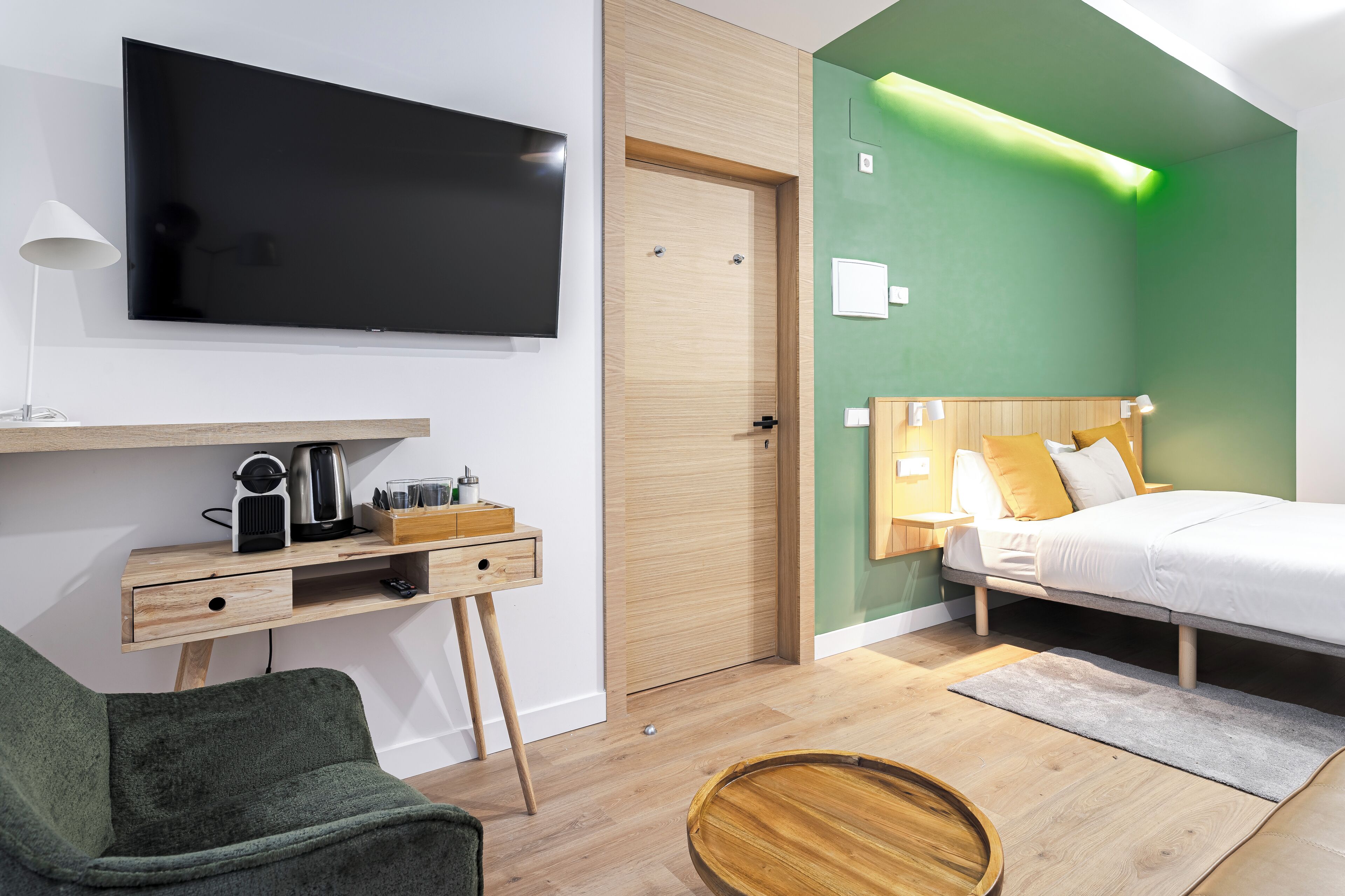 Una habitación de hotel moderna con televisor de pantalla plana, escritorio de madera, paredes verdes y una cama con ropa blanca.