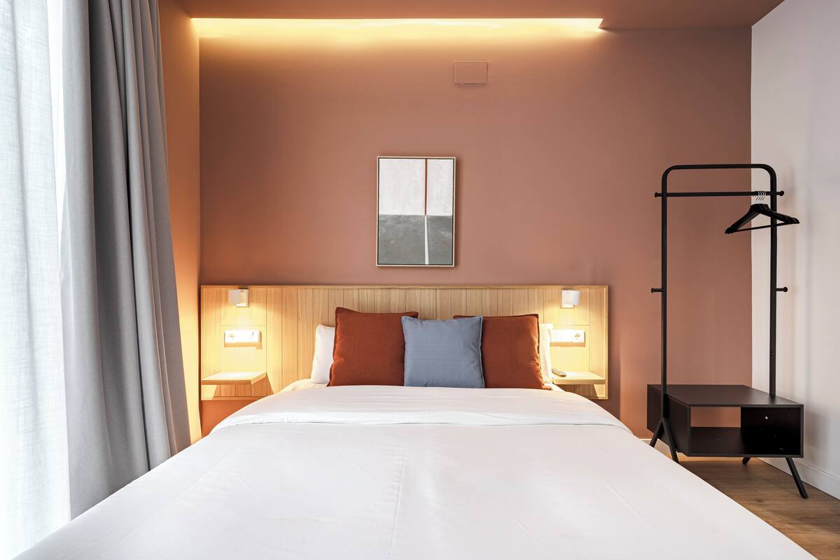 Une chambre d’hôtel avec un grand lit, deux tables de chevet avec des lampes, une tête de lit en bois, une œuvre d’art accrochée au mur et un porte-manteau noir.