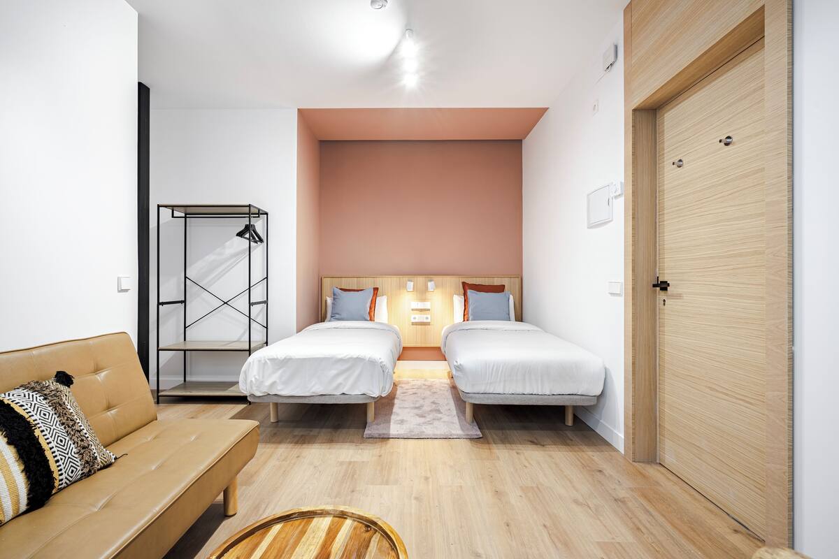 Une chambre d’hôtel moderne avec deux lits simples, un plancher en bois, un canapé et une armoire.