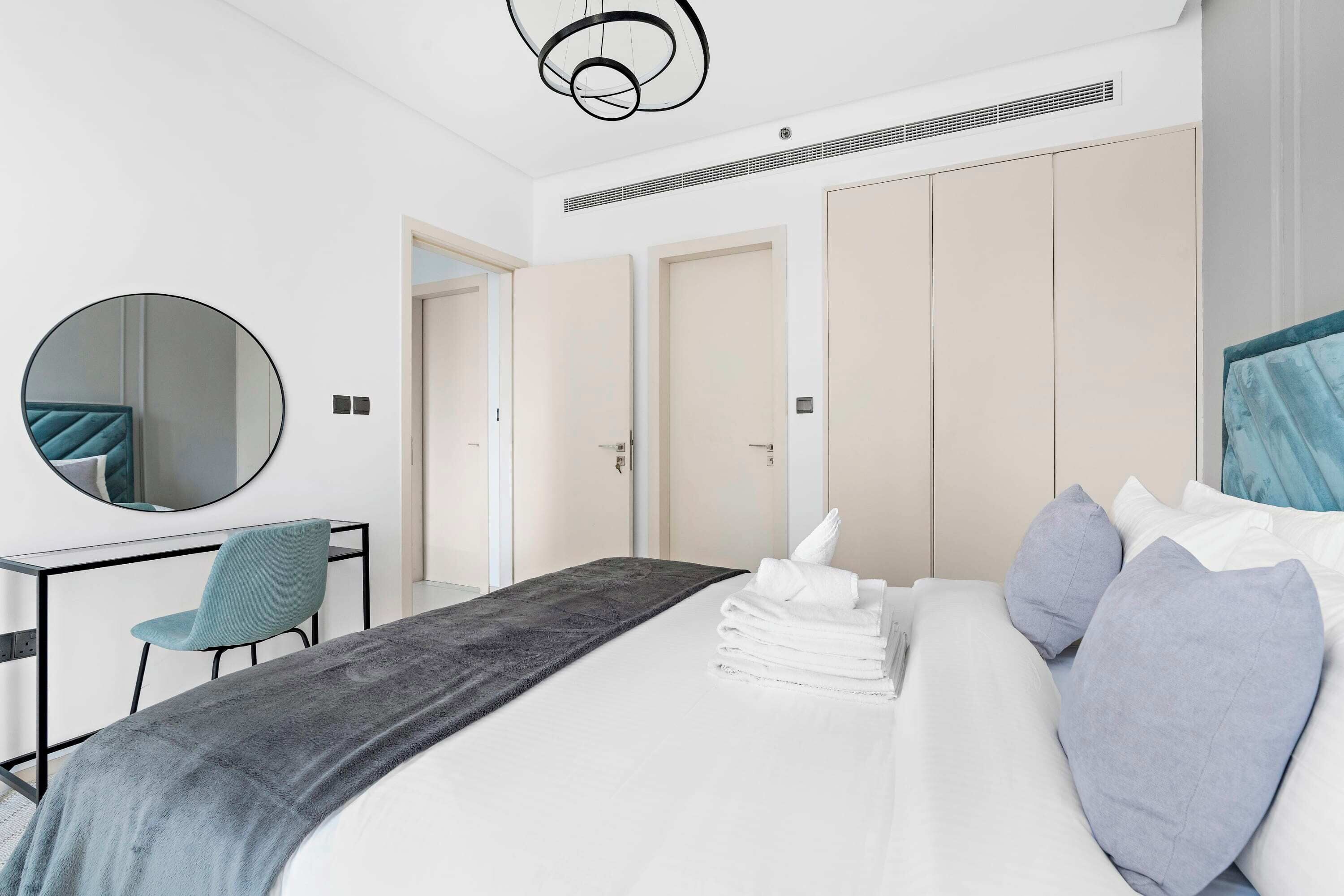 A modern hotel room with a bed, a desk, a chair, a round mirror, and a wardrobe.