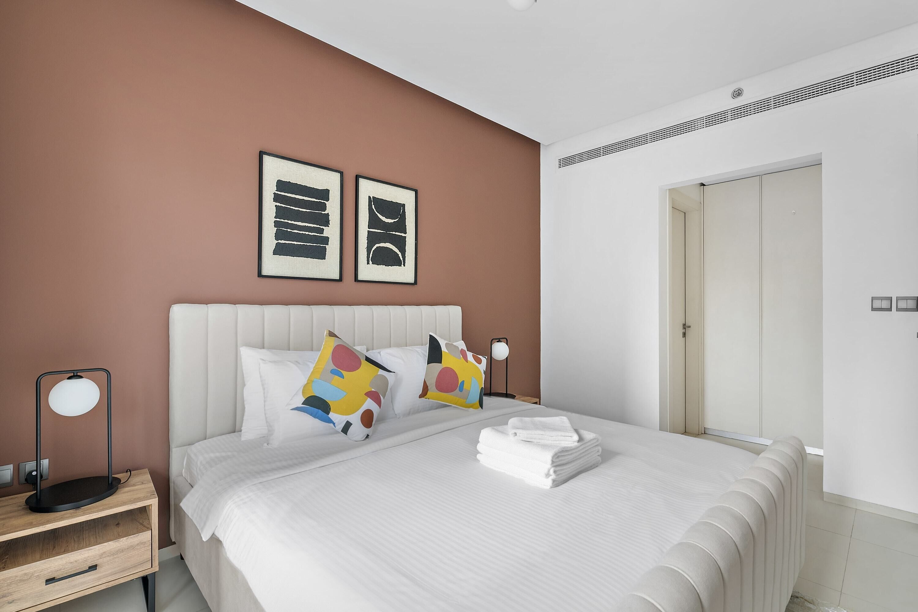 A modern hotel room with a large bed, white bedding, and colorful pillows. There is a bedside table with a lamp and a small drawer. The room has a brown accent wall and white walls.