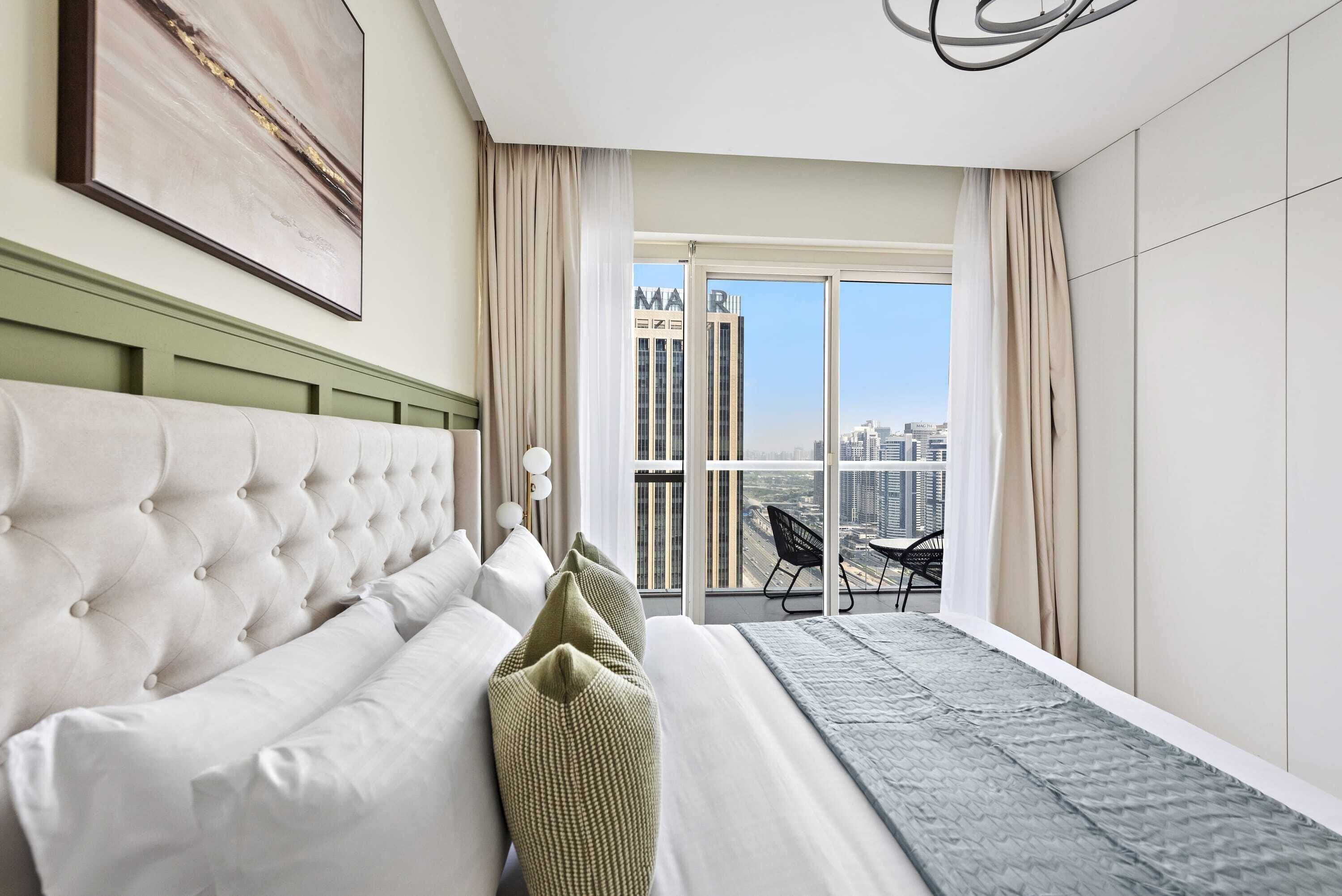 A modern hotel room with a large bed, a view of the city, and a balcony with a table and chairs.
