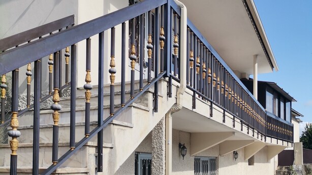A two-story house with a stone facade, a balcony with ornate balustrades, and a cobblestone pathway.
