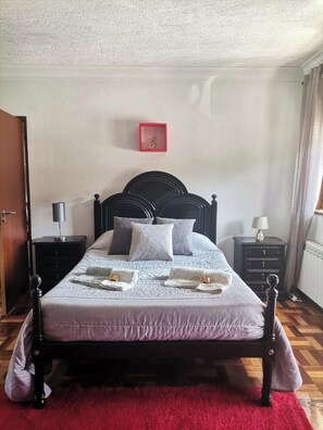 Een net opgemaakt bed met hoofdbord, een foto met rode lijst aan de muur, en een rood vloerkleed.