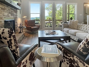 A living room with a stone fireplace, leather armchairs, a coffee table, and a view through large glass doors. - Mountain-view cabin with hot tub, game room, and 3 bedrooms. Free rafting, train, zip (Bryson City)