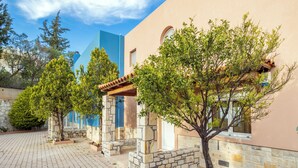 A courtyard with stone paving, trees, and a building with a stone facade and blue accents. - Nami Villa SeaView | Private Pool  (Heraklion)
