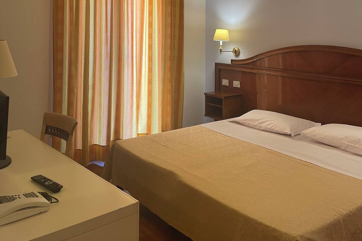 Habitació d'hotel amb llit, escriptori amb làmpada, cadira i finestra amb cortines.