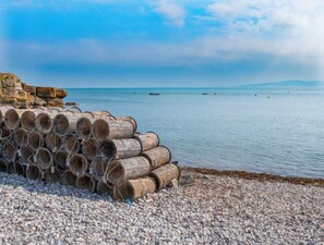 A coastal scene with a stack of cylindrical logs on a pebbly beach, overlooking calm sea and distant rocky cliffs. - Heulwen - sleeps 7 - Moelfre - Anglesey (Moelfe)