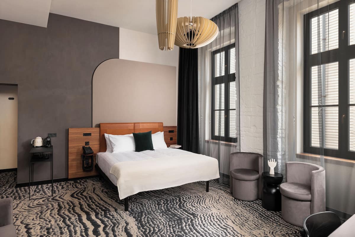 Una habitación de hotel moderna con una cama grande, un cabecero de madera, una alfombra estampada y vistas a la ciudad a través de amplios ventanales.