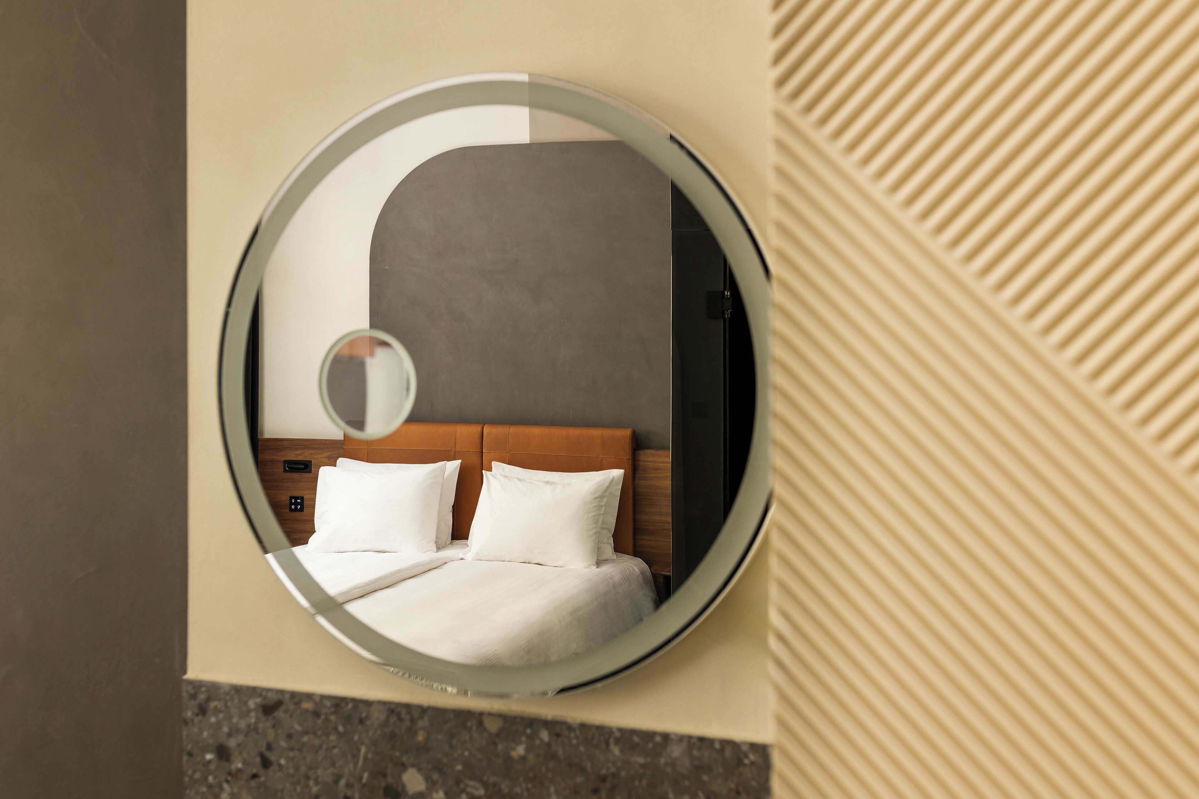 A circular mirror reflecting a neatly made bed with white bedding and a wooden headboard.