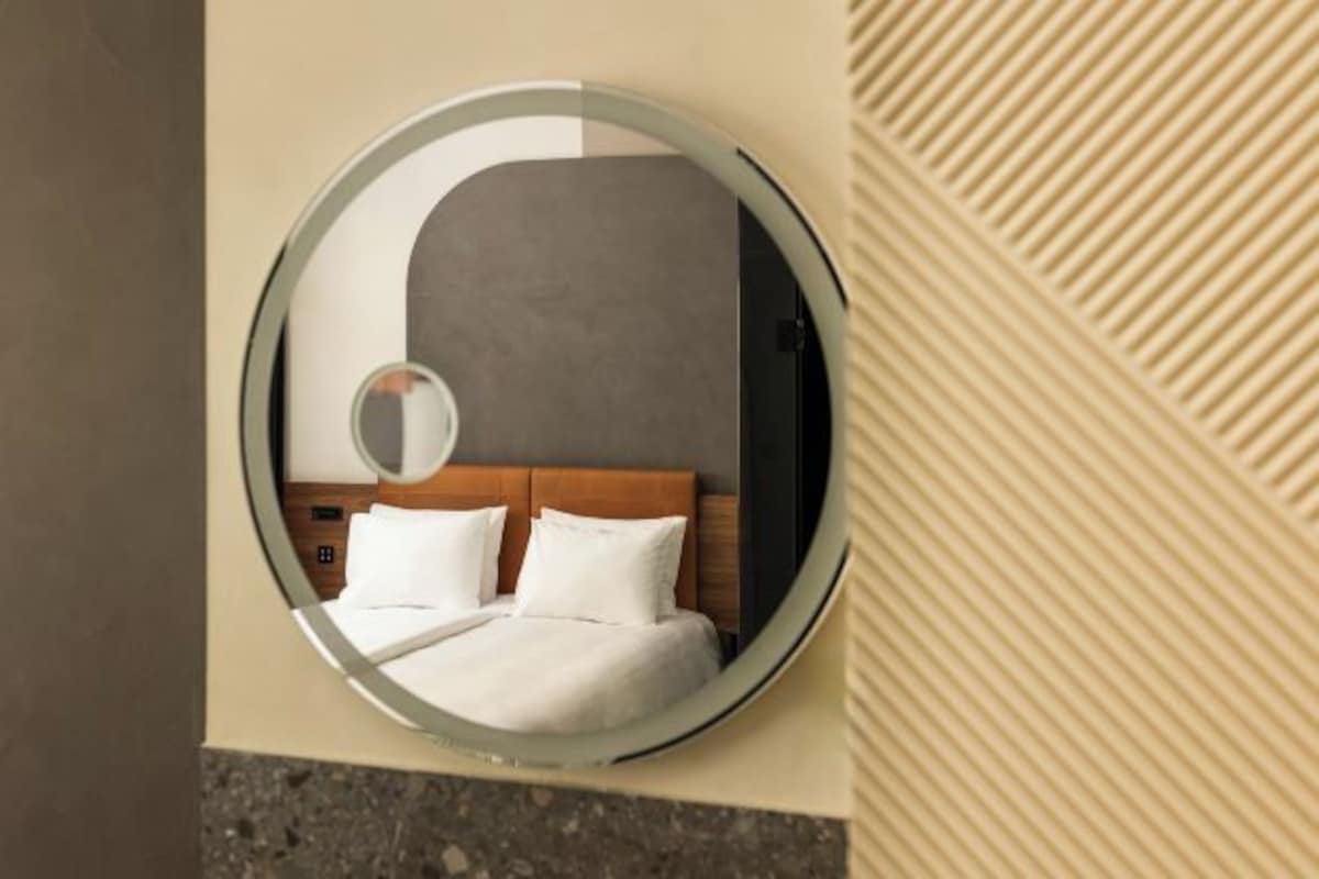 Un espejo circular que refleja una cama con ropa de cama blanca y un cabecero de madera.