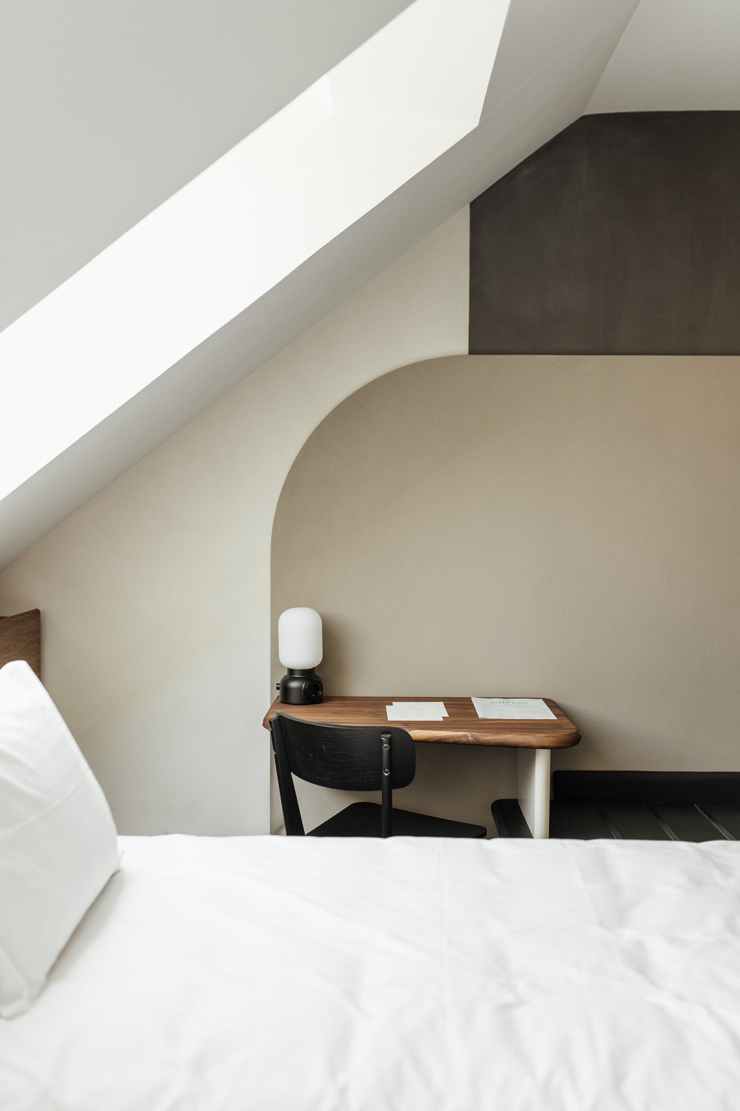 A bedroom with a sloped ceiling, a bed, a desk with a chair, and a wall-mounted lamp.