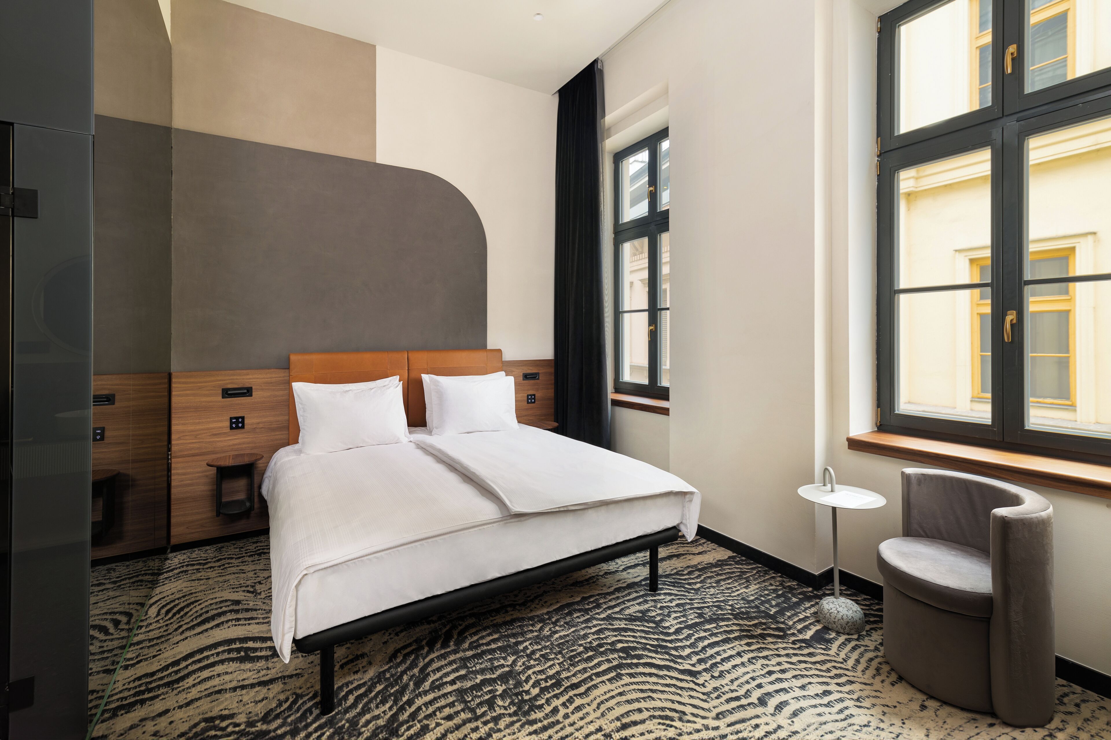 A hotel room with a bed, a small round table, a chair, and a window with a view of buildings.