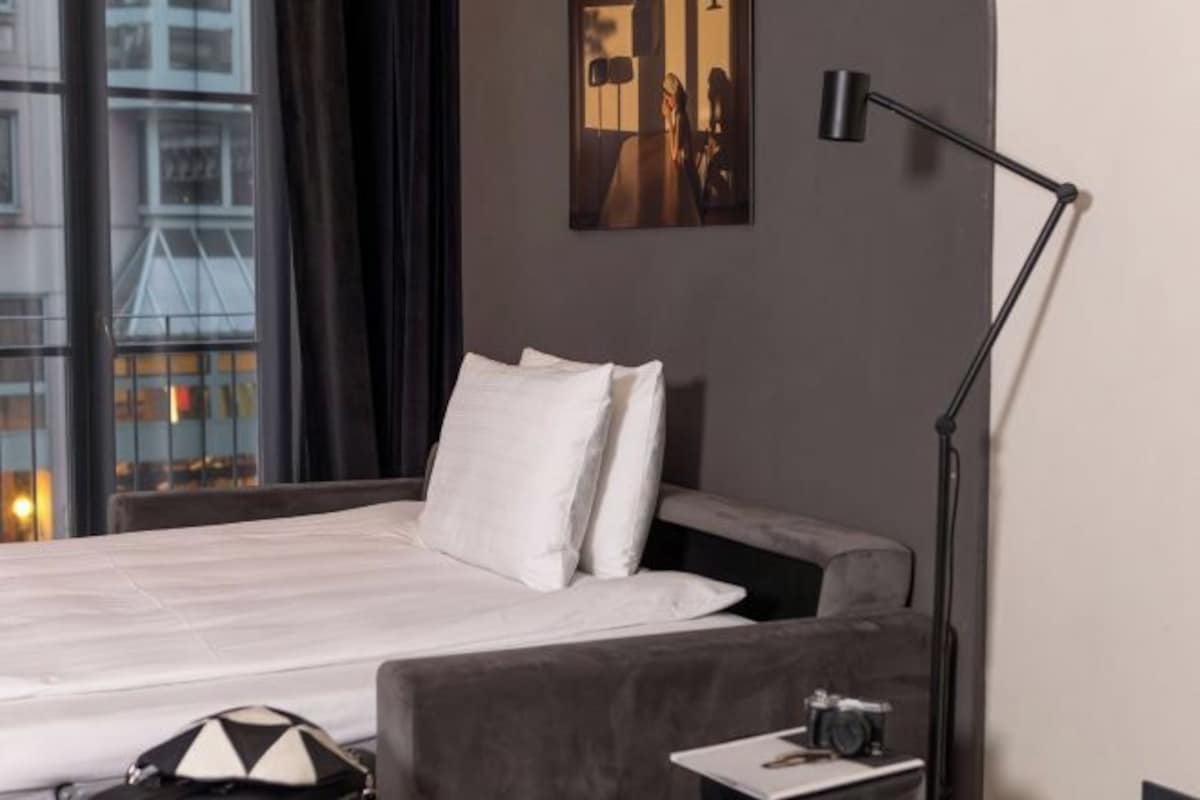 Habitación de hotel con cama, mesita de noche, lámpara y ventana con vistas a edificios.
