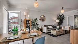 Een moderne woonkamer met een eethoek, met een houten eettafel, blauwe stoelen, een bank en een salontafel. Er staan twee potplanten, een houten kast en een grote spiegel aan de muur.