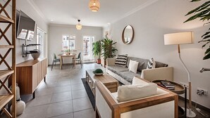 Een moderne woonkamer met een bank, salontafel en een eethoek met stoelen. Er is een televisie aan de muur bevestigd, een plant en een lamp.