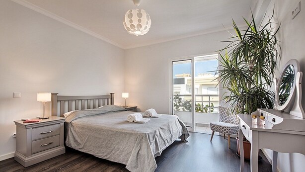 Een slaapkamer met een bed, nachtkastjes, een bureau met spiegel, een potplant en een balkon met uitzicht.
